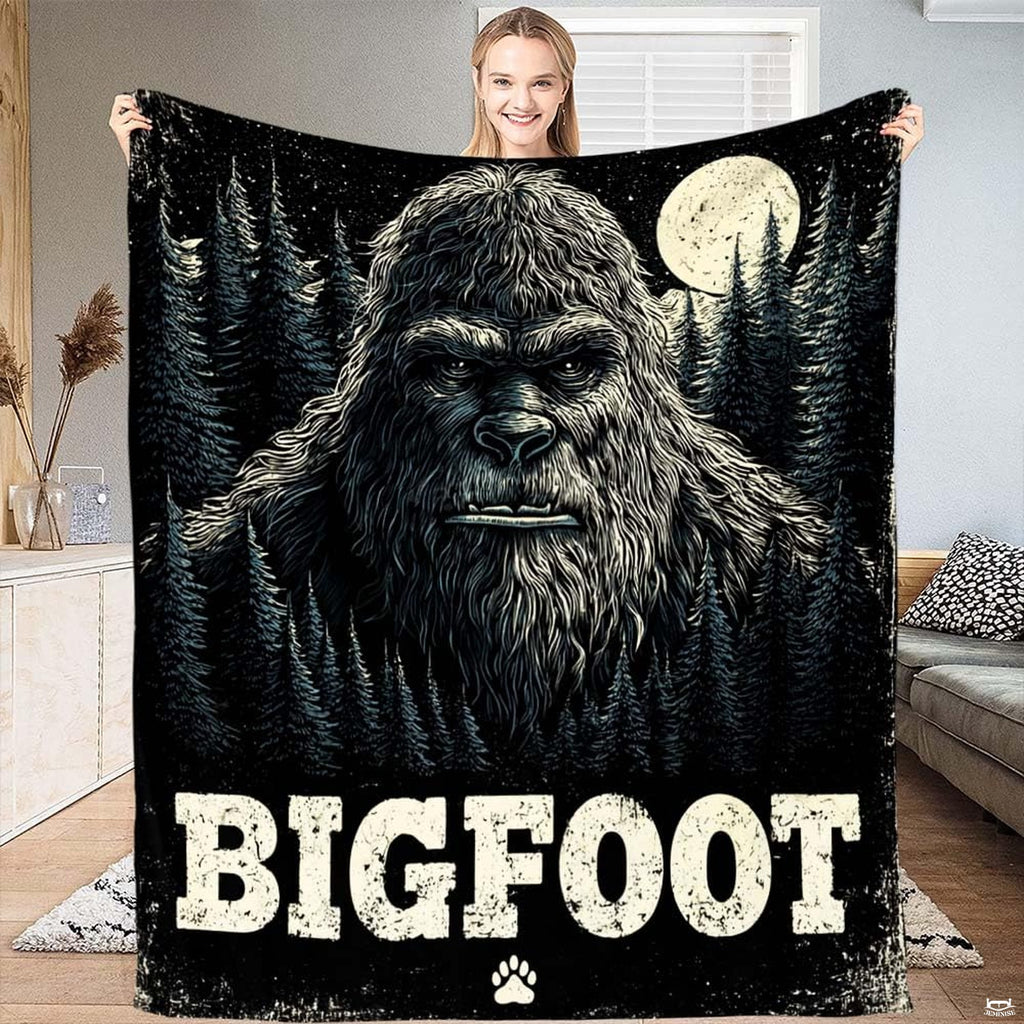 Sasquatch face illustration against a full moon, stars, and evergreen pines on a decorative throw blanket