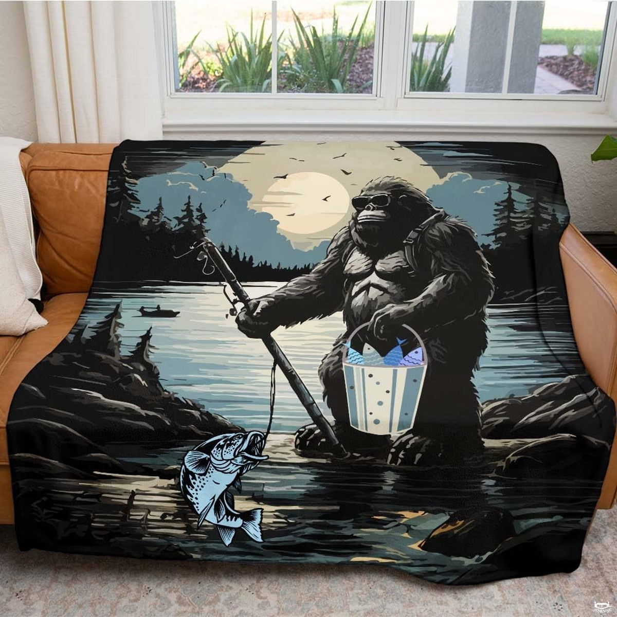 Fishing Bigfoot graphic throw blanket draped over brown leather living room sofa with lake scene.