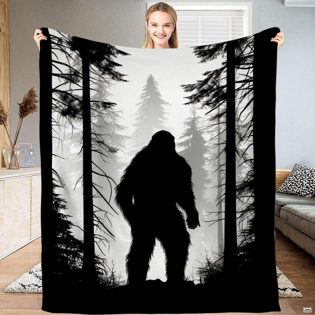 Woman holding up a monochrome blanket displaying a Bigfoot silhouette standing in a misty pine forest.