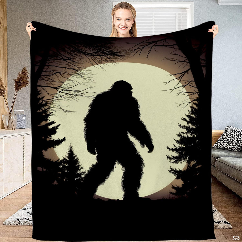 Woman holding up a black blanket displaying a Bigfoot silhouette walking past pines before an ivory moon.