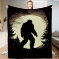 Woman holding up a black blanket displaying a Bigfoot silhouette walking past pines before an ivory moon.