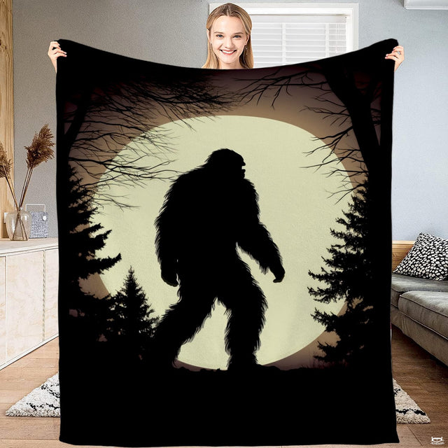 Woman holding up a black blanket displaying a Bigfoot silhouette walking past pines before an ivory moon.