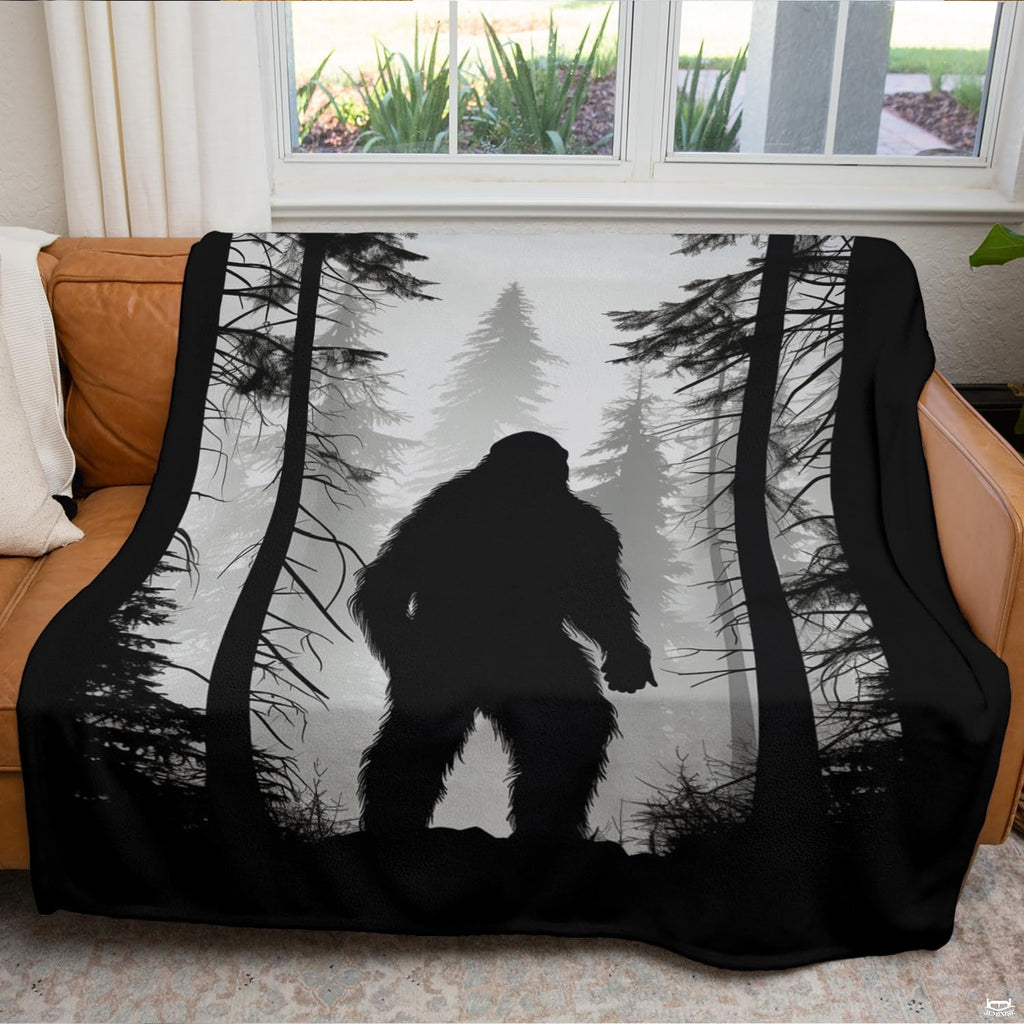 Misty pine forest Bigfoot blanket draped across an indoor living room couch near a window.