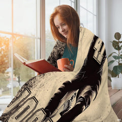 Person wrapped comfortably in sports throw blanket while reading a book beside a sunny window.