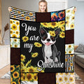 French Bulldog puppy wearing floral crown surrounded by bright yellow sunflower blossoms on bedding.