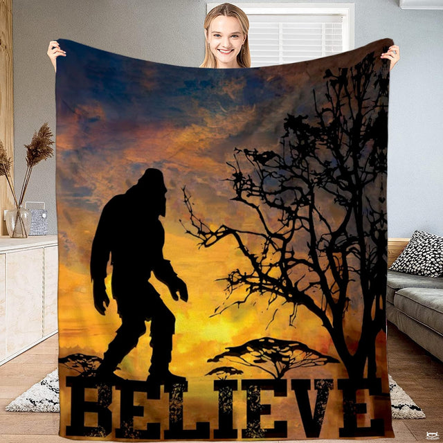 Bigfoot blanket held open showing walking Sasquatch silhouette against a fiery sunset and gnarled tree.