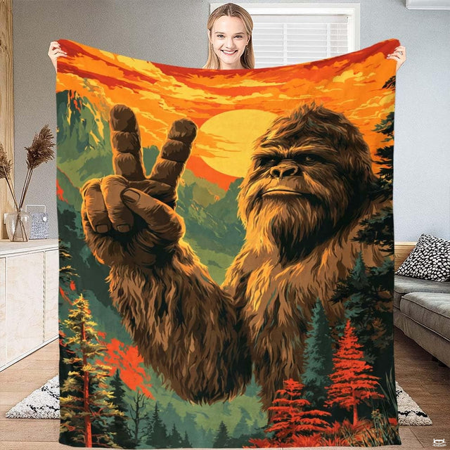 Woman holding up a vibrant blanket displaying Bigfoot flashing a peace sign before a mountain sunset.