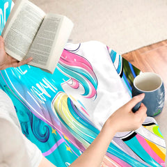 Person sitting with book and warm mug while covered in colorful unicorn graphic throw blanket.