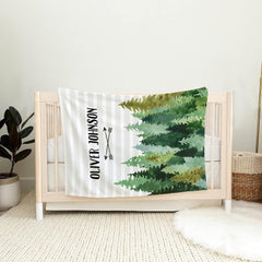 Nursery scene showing woodland pine tree graphic draped over a light wooden crib railing beside a plant.