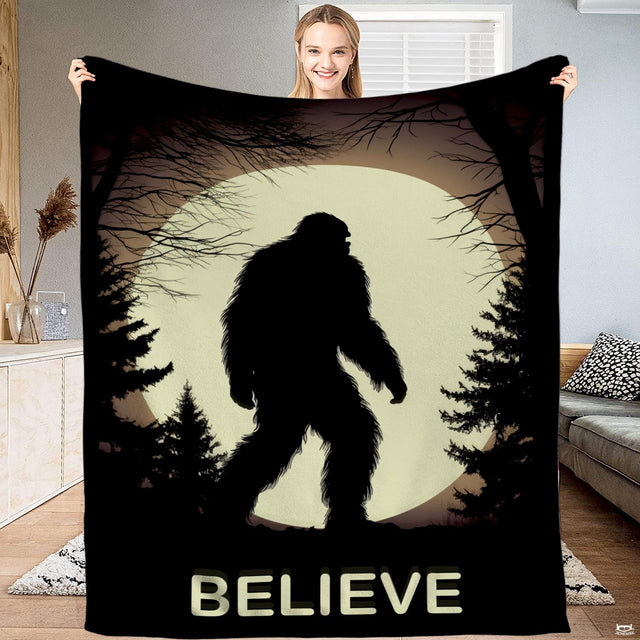Woman holding up a black blanket displaying a Bigfoot silhouette walking past pines before a circular moon.