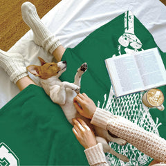 Puppy resting on a draped emerald sports blanket beside an open book and beverage mug.