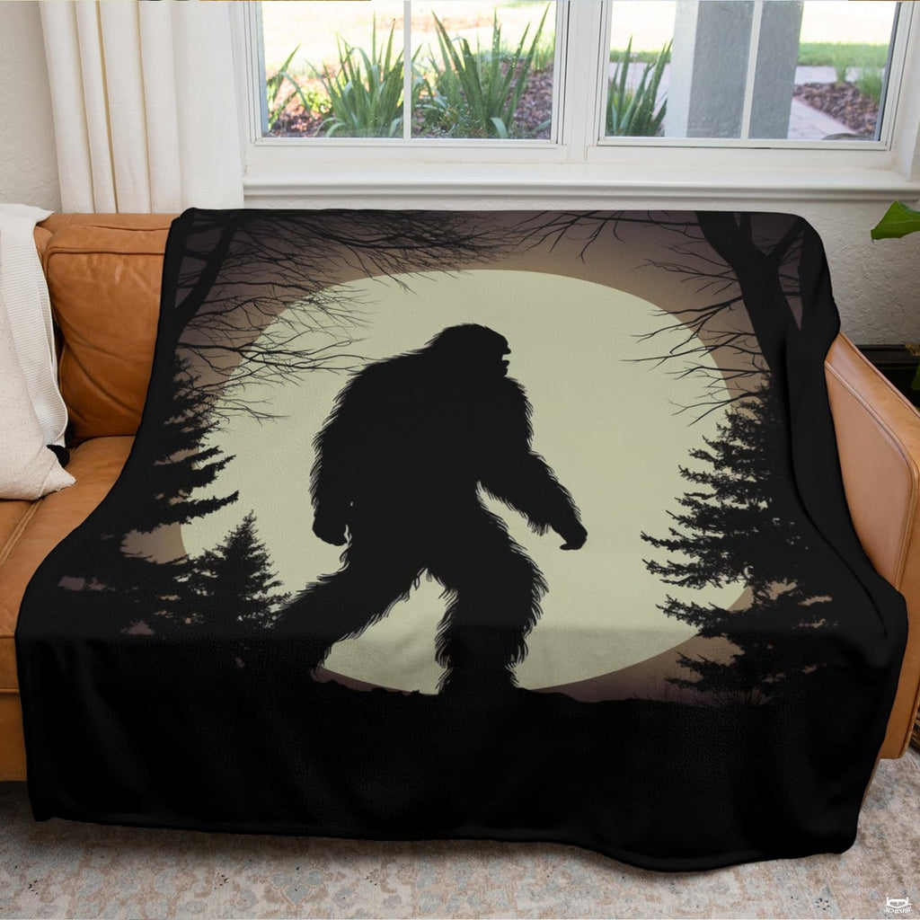 Pale moon Bigfoot forest blanket draped across an indoor living room couch near a bright window.