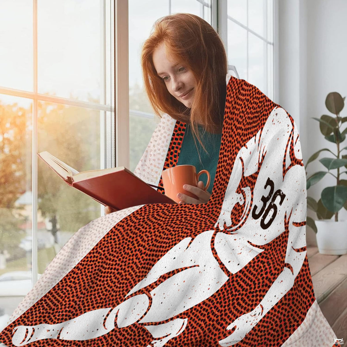 Woman wrapped comfortably in football throw blanket while reading a book near a sunny window.